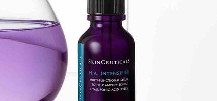 Een paarse fles huidserum van SkinCeuticals met een pipet, op een lichte achtergrond naast een glazen watercontainer.