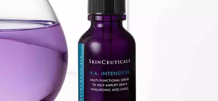 Een paarse fles huidserum van SkinCeuticals met een pipet, op een lichte achtergrond naast een glazen watercontainer.