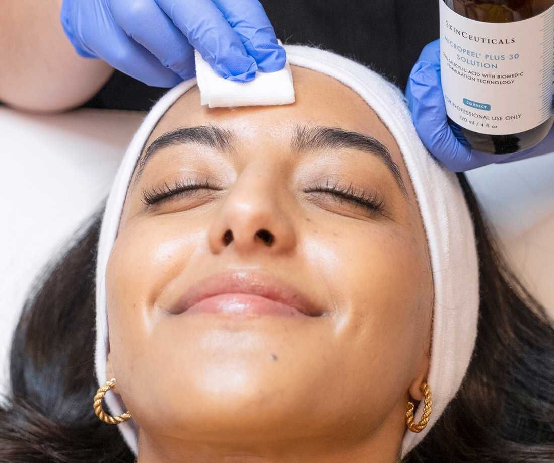Une femme portant un bandeau blanc est allongee sur une table de traitement, tandis qu'une personne avec des gants bleus applique un soin du visage a l'aide d'un coton.