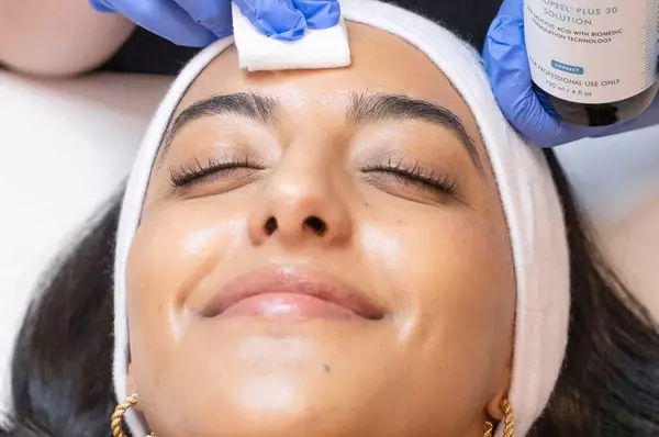 Une femme portant un bandeau blanc est allongee sur une table de traitement, tandis qu'une personne avec des gants bleus applique un soin du visage a l'aide d'un coton.