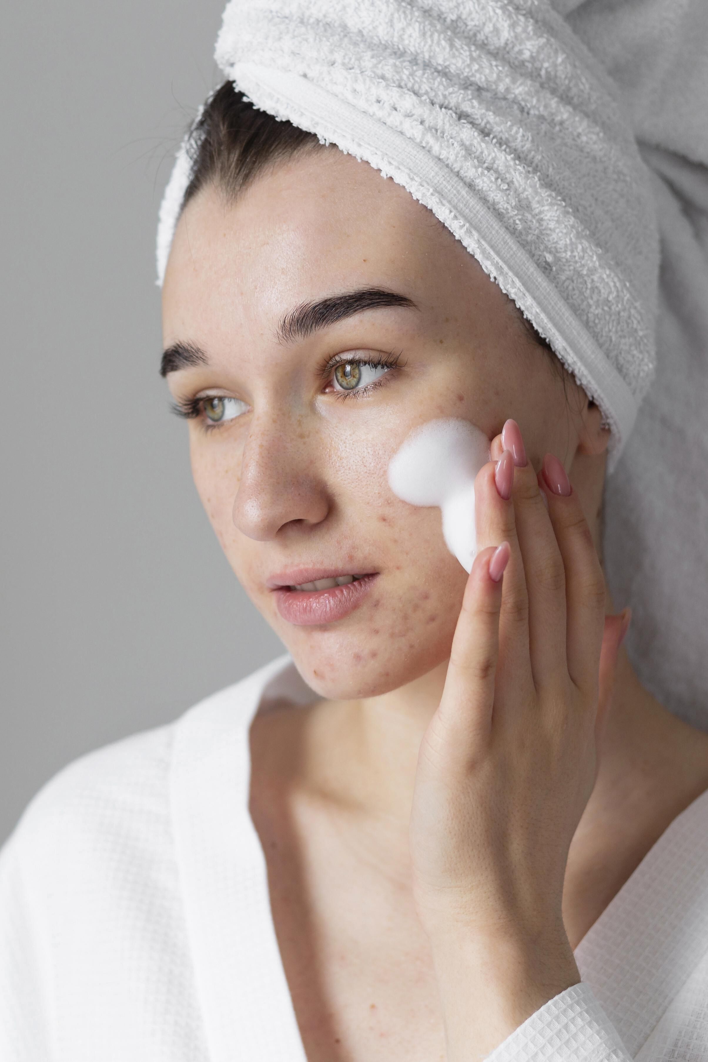 Une femme en serviette de bain appliquant un nettoyant moussant sur son visage, avec un aspect naturel et frais.