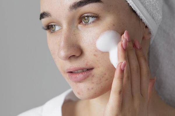Une femme en serviette de bain appliquant un nettoyant moussant sur son visage, avec un aspect naturel et frais.
