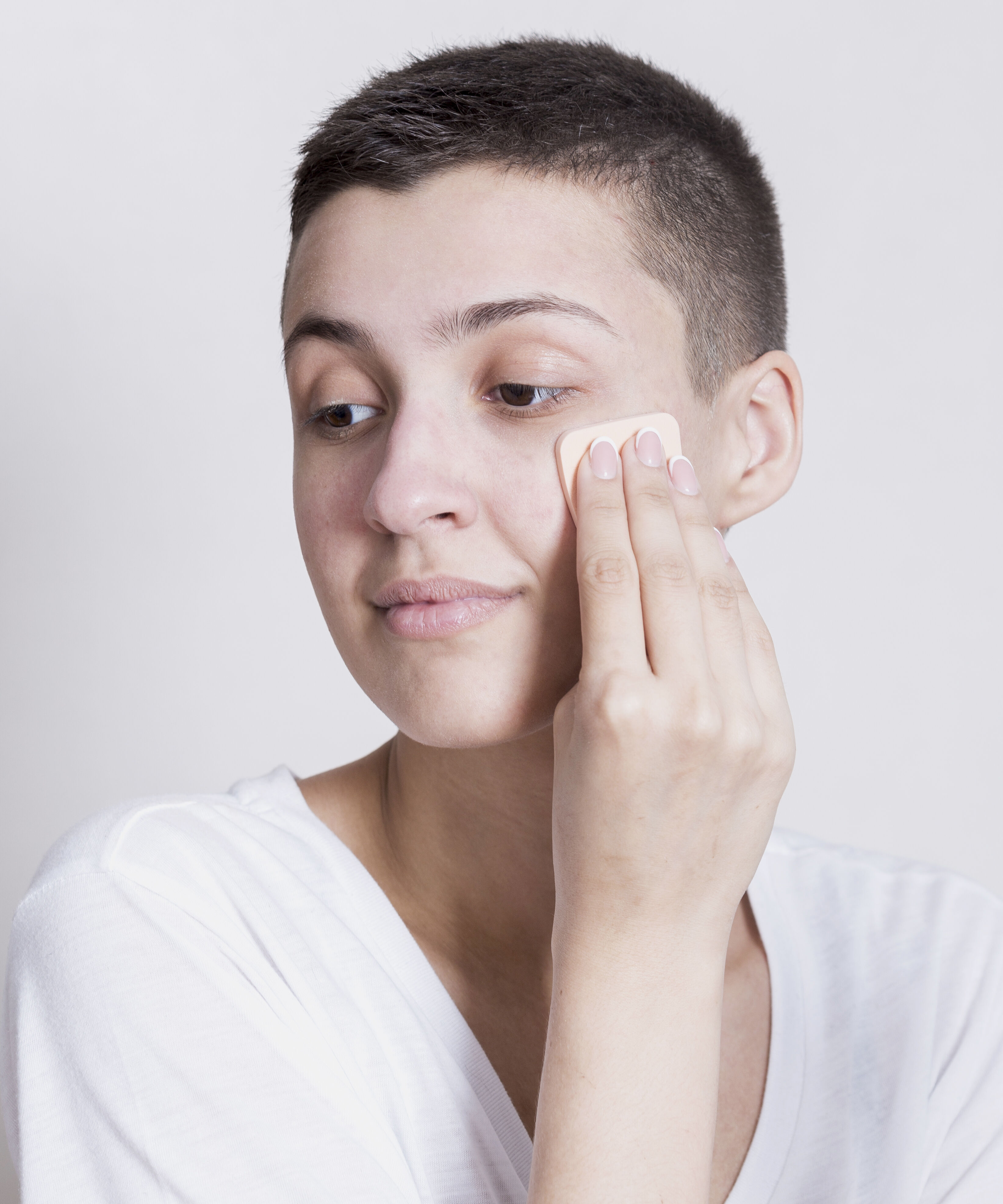 Une personne aux cheveux courts applique du fond de teint sur son visage a l'aide d'une eponge de maquillage.