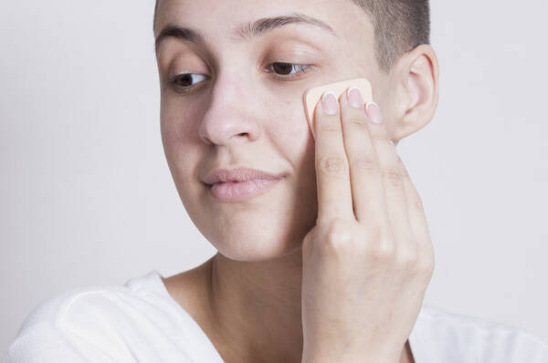 Une personne aux cheveux courts applique du fond de teint sur son visage a l'aide d'une eponge de maquillage.