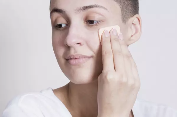 Une personne aux cheveux courts applique du fond de teint sur son visage a l'aide d'une eponge de maquillage.