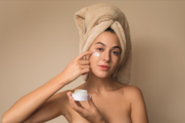 Une femme avec une serviette autour de la tete applique une creme pour le visage sur son visage. Elle tient un pot de creme dans son autre main et regarde directement la camera. L'arriere-plan est de couleur neutre.