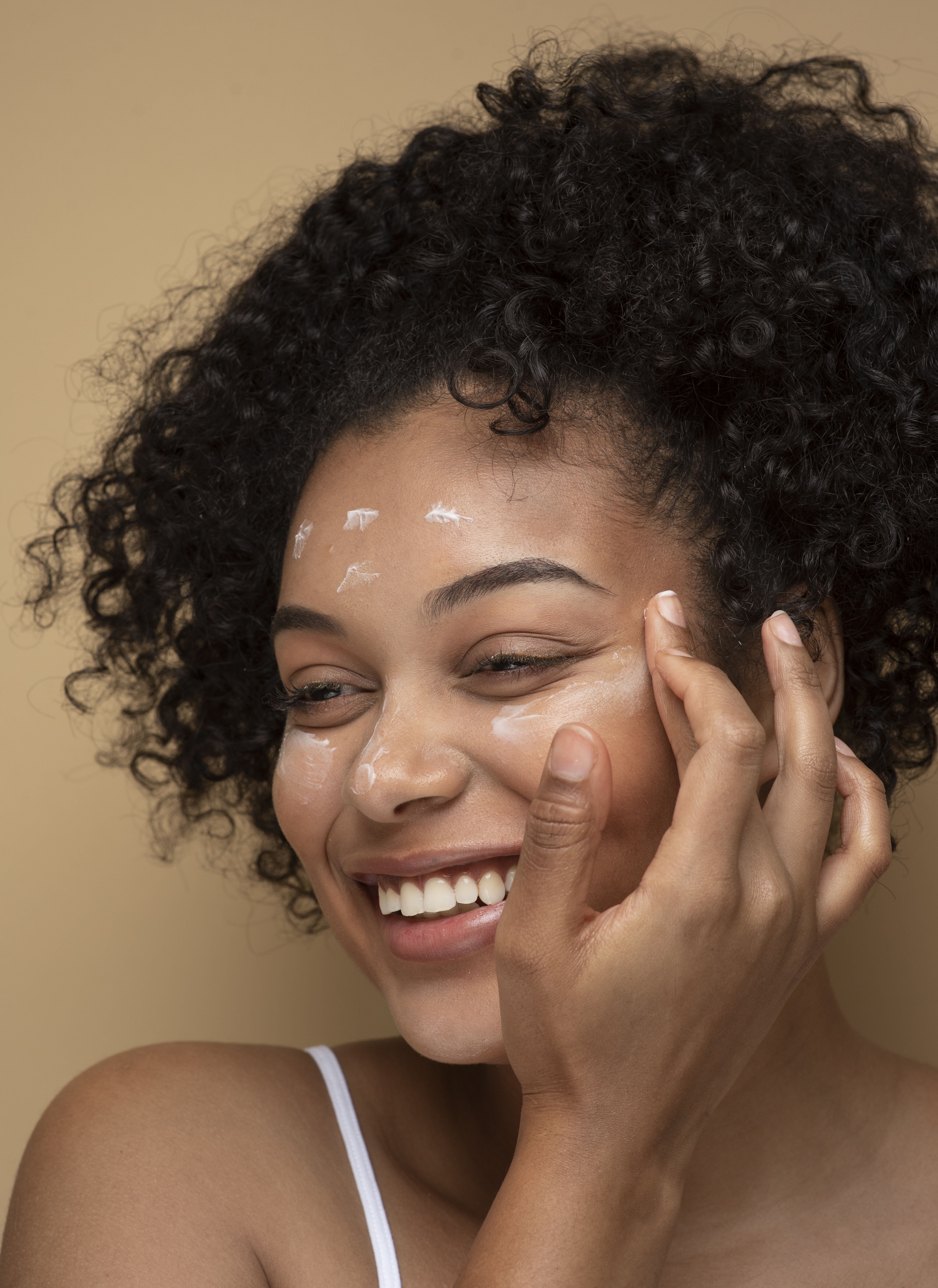Une femme aux cheveux boucles, souriant en appliquant une creme sur son visage, sur un fond beige.