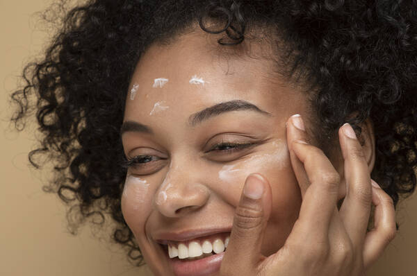 Une femme aux cheveux boucles, souriant en appliquant une creme sur son visage, sur un fond beige.