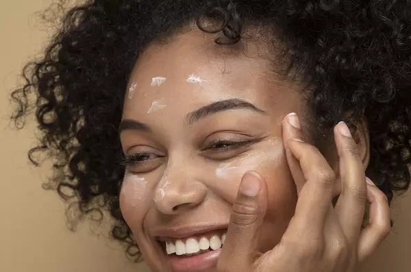 Une femme aux cheveux boucles, souriant en appliquant une creme sur son visage, sur un fond beige.