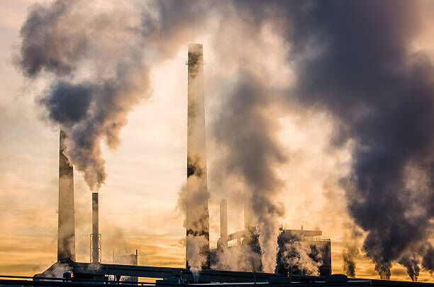 Rookpluimen stijgen op uit hoge schoorstenen van een industriegebied tegen een zonsopgang.