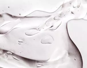 Close-up of clear gel-like substance with smooth curves and small, glistening droplets on a light background.