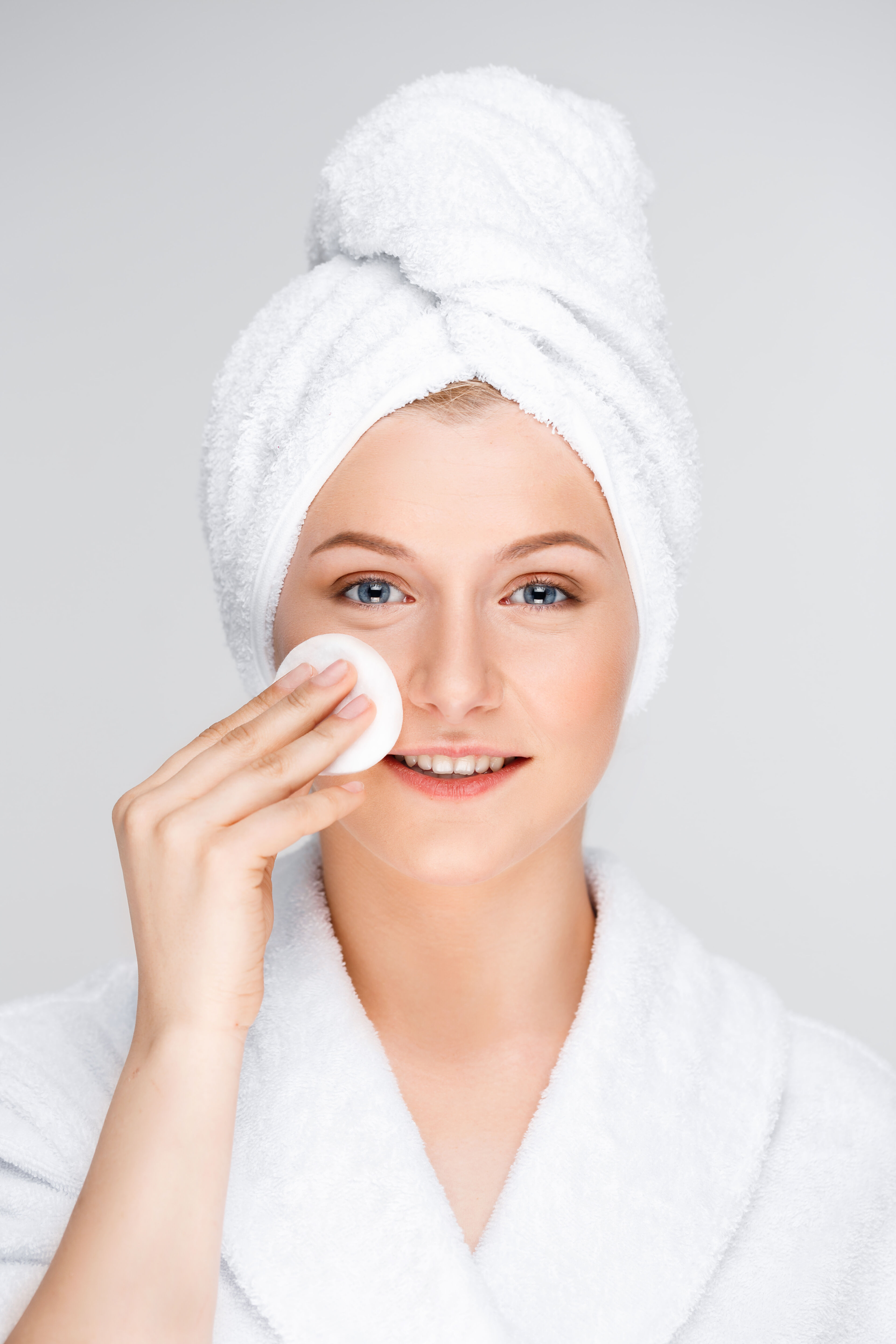 Une femme en peignoir blanc et avec une serviette autour de la tete, appliquant un coton sur son visage.