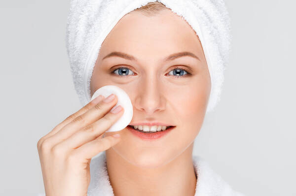 Une femme en peignoir blanc et avec une serviette autour de la tete, appliquant un coton sur son visage.
