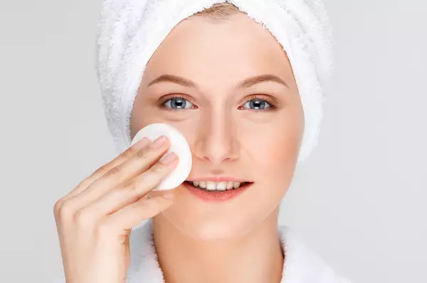 Une femme en peignoir blanc et avec une serviette autour de la tete, appliquant un coton sur son visage.