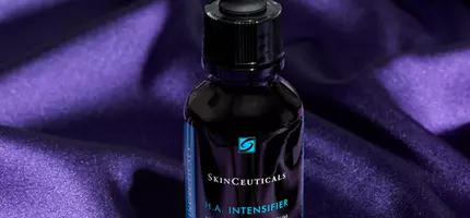 A bottle of SkinCeuticals H.A. Intensifier serum sitting on a purple satin background, featuring a dropper top and a sleek design.