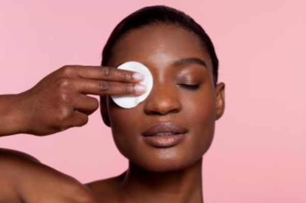 Une femme appliquant un coton avec nettoyant visage sur son visage, avec un arriere-plan rose doux.