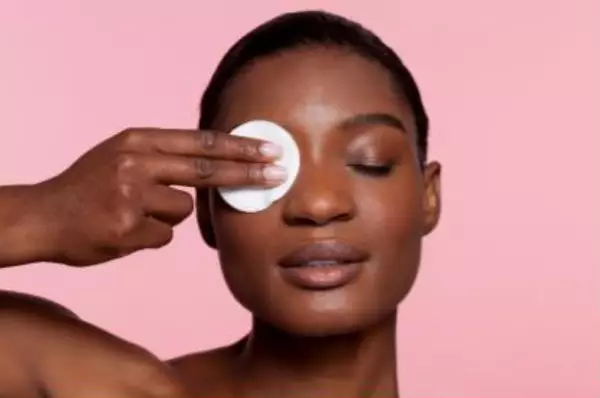 Une femme appliquant un coton avec nettoyant visage sur son visage, avec un arriere-plan rose doux.