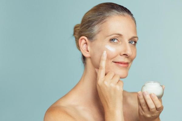 Femme torse nu souriante appliquant une creme sur sa joue tout en tenant un pot de creme.