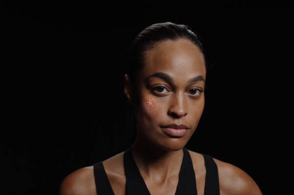 Une femme regarde directement la camera, avec une expression neutre sur son visage et un dessin subtil sur sa joue, sur un fond noir.