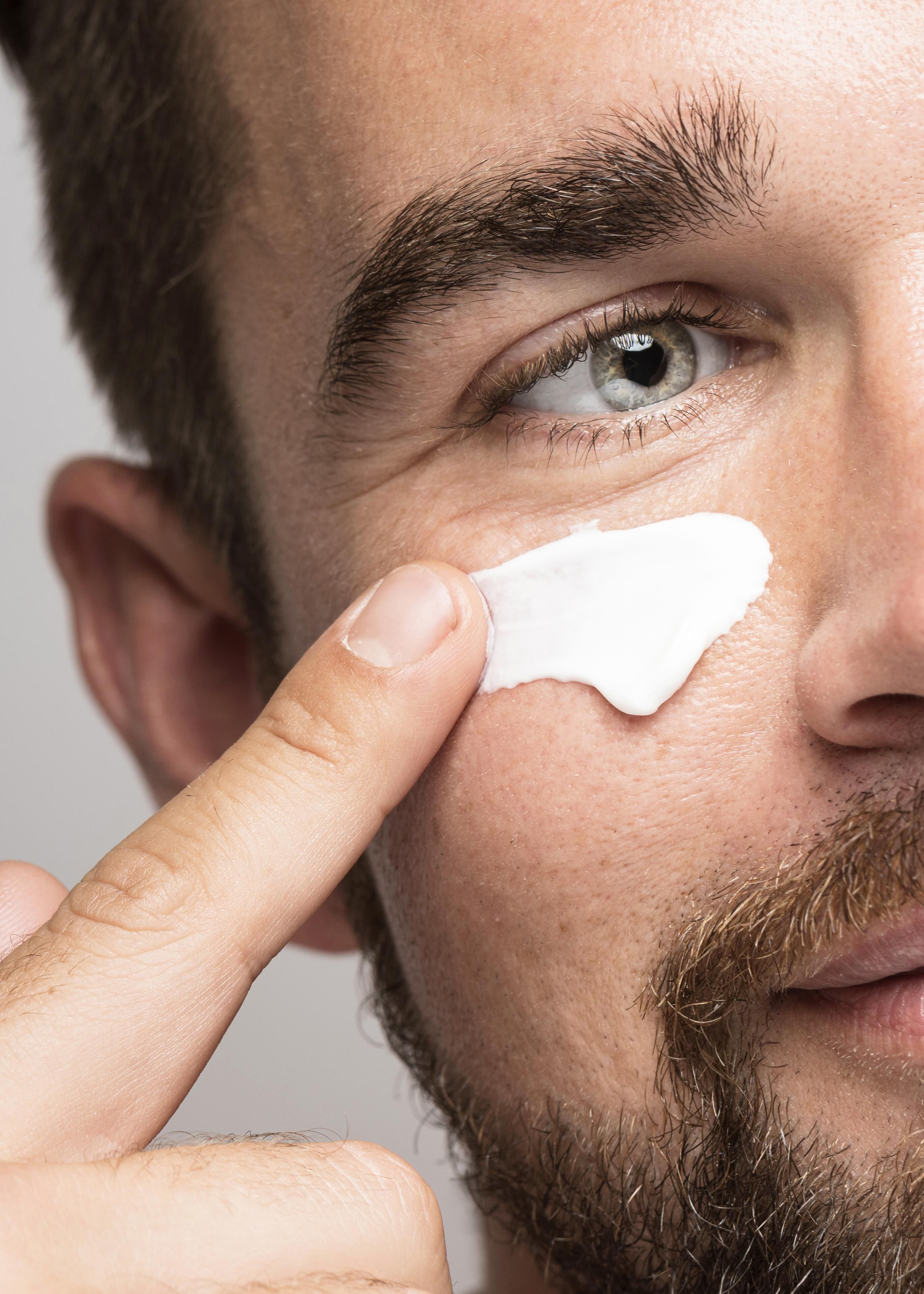 Un homme barbu appliquant une creme visage sur sa peau, tout en pointant du doigt le produit.