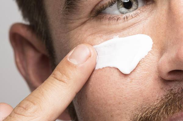 Un homme barbu appliquant une creme visage sur sa peau, tout en pointant du doigt le produit.
