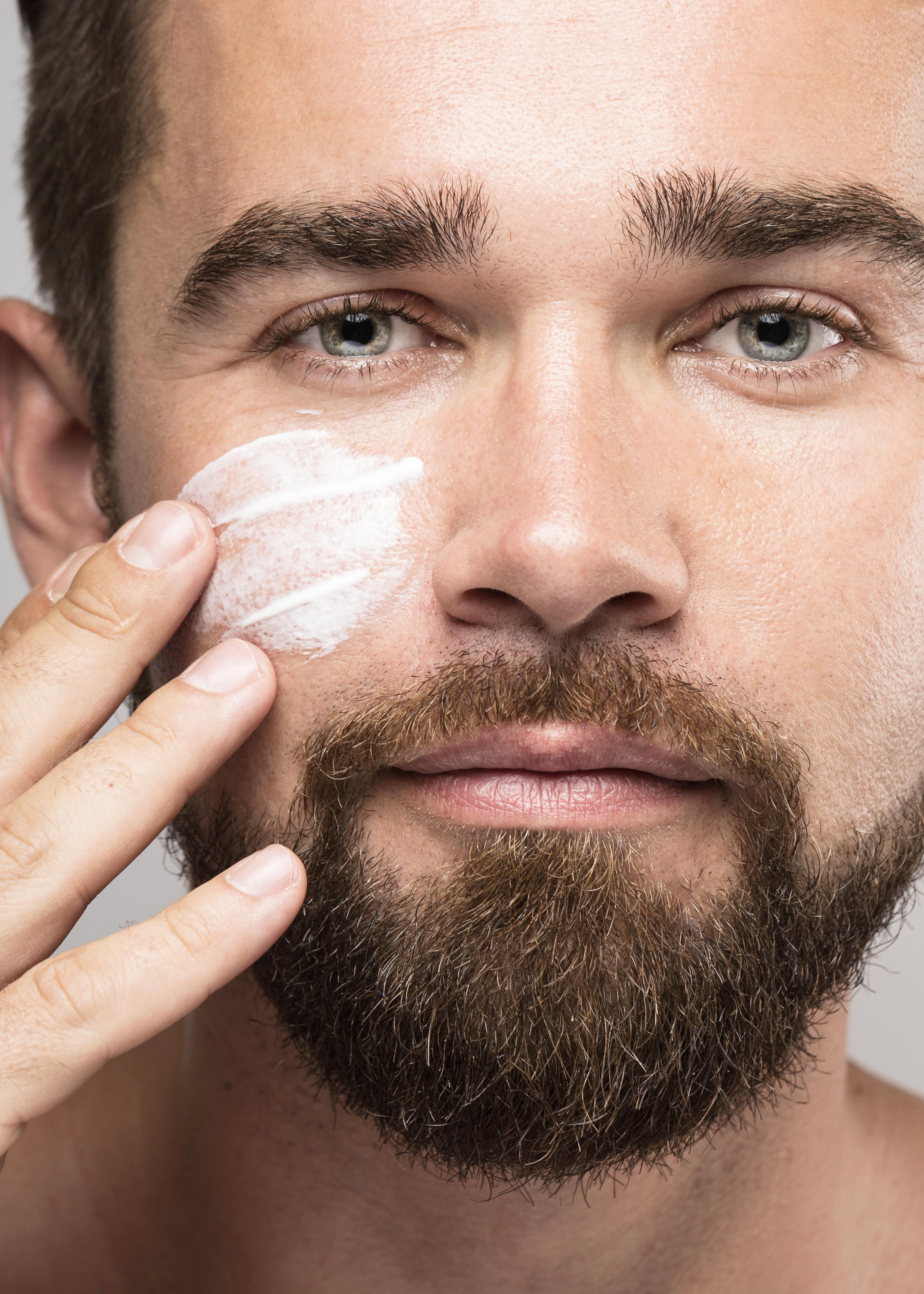 Un homme avec une barbe applique une creme visage sur son visage.