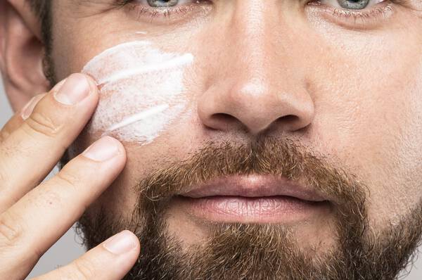 Un homme avec une barbe applique une creme visage sur son visage.