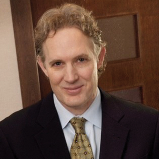 A smiling man in a suit, standing in an office setting with wooden doors in the background.